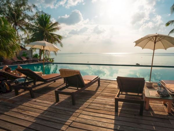 Poolside scene with lounge chairs, umbrellas, palm trees, and a serene ocean view under a partly cloudy sky.