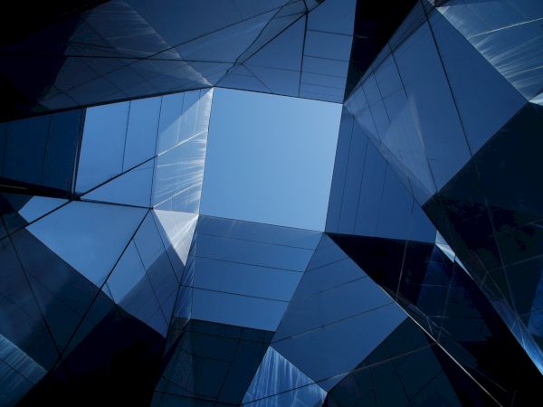 Geometric view of a modern glass building with reflective surfaces, creating an abstract pattern framing the sky in the center.