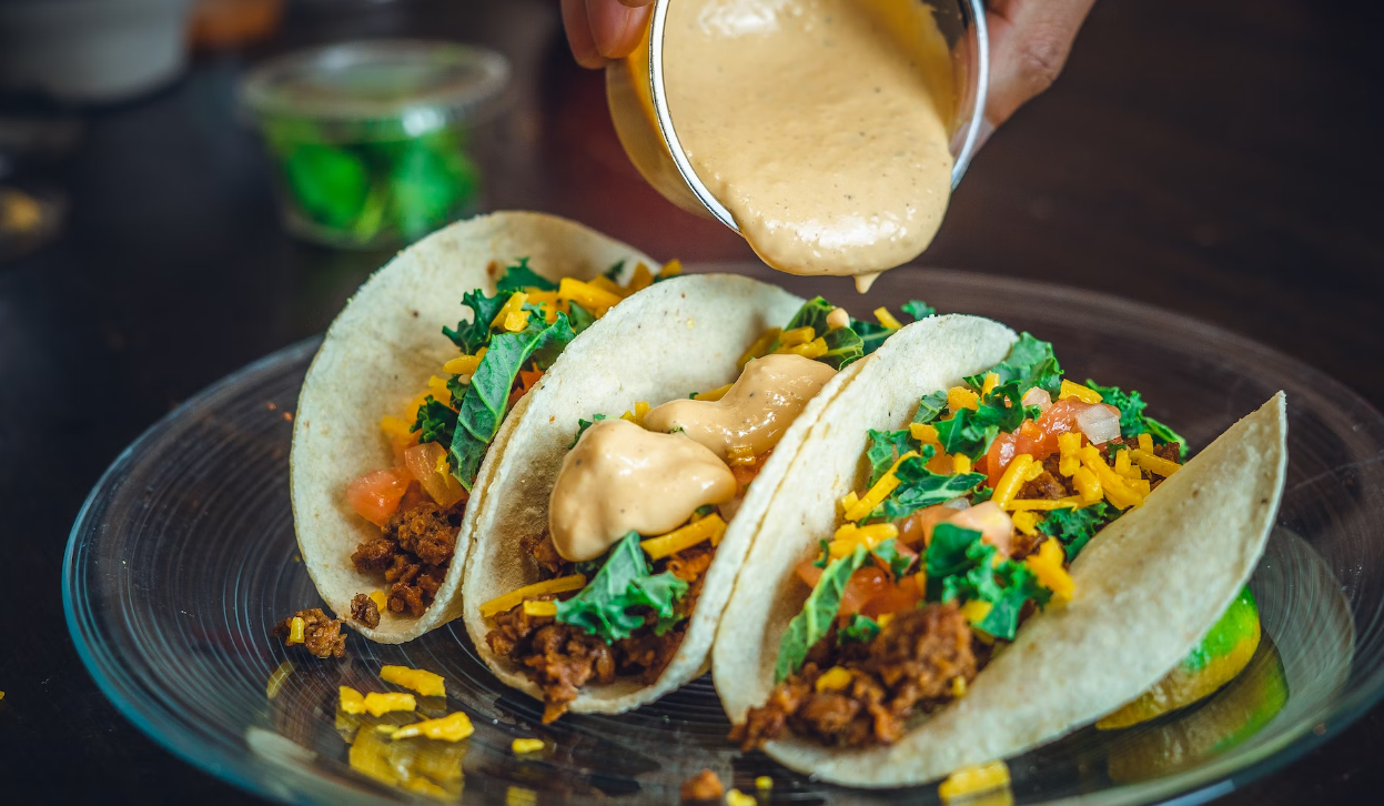 Three tacos filled with ground meat, cheese, lettuce, and tomatoes on a plate, being topped with a creamy sauce.