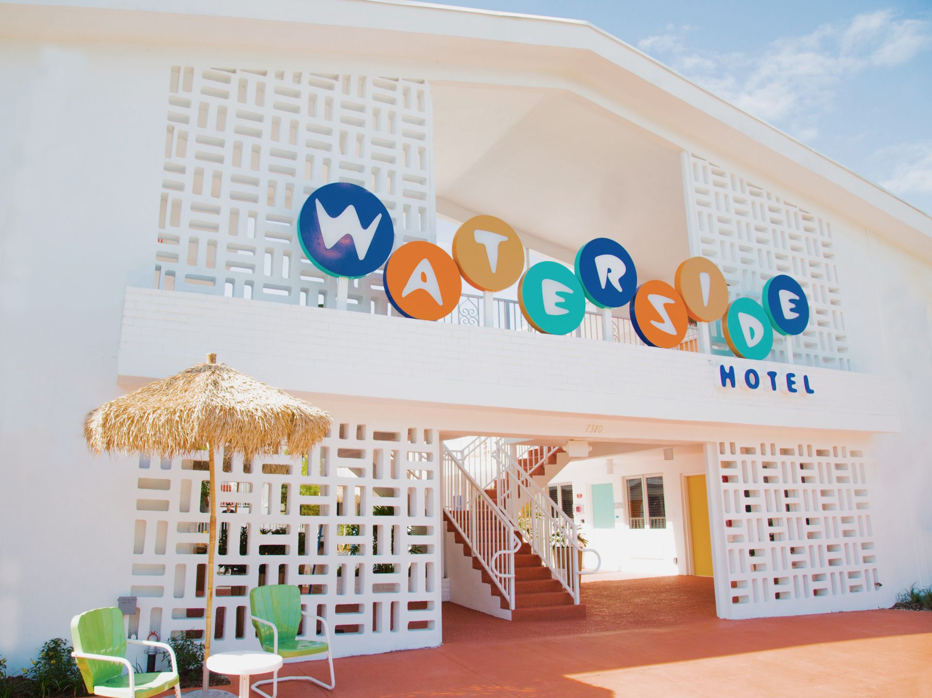 The image shows the entrance of a hotel with colorful "Waterside Hotel" signage, decorative white walls, chairs, and a small thatched umbrella.