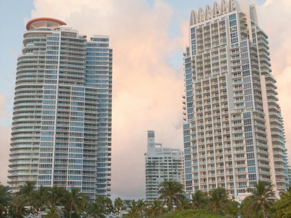 Two modern high-rise buildings, with a sandy beach and palm trees in the foreground, under a sky with cotton candy-like clouds.