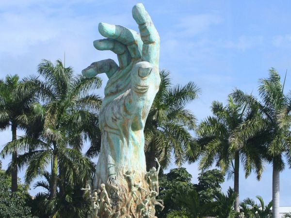 A large sculpture of a hand reaching upward, surrounded by small human figures, set against a backdrop of palm trees and blue sky.