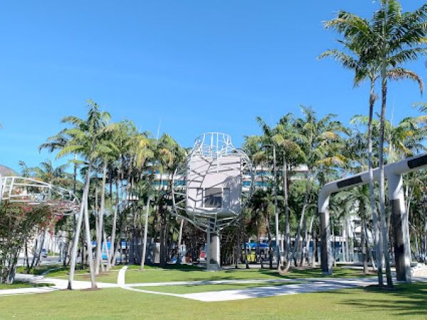 A park with palm trees, grass, and modern metal structures under a clear blue sky.