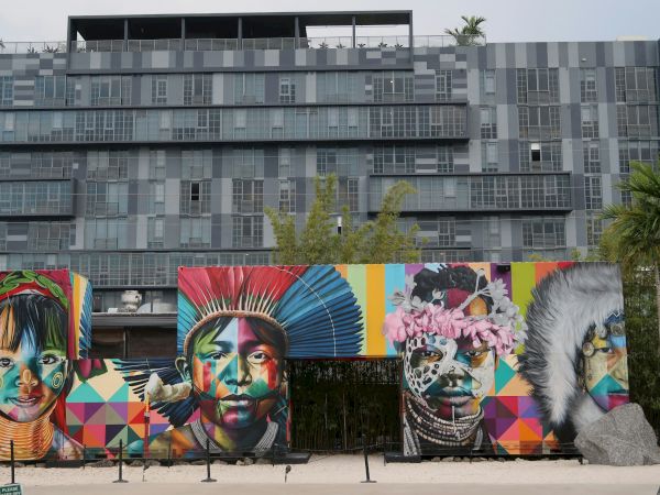The image shows colorful murals of diverse faces on a building, with geometric patterns and greenery around.