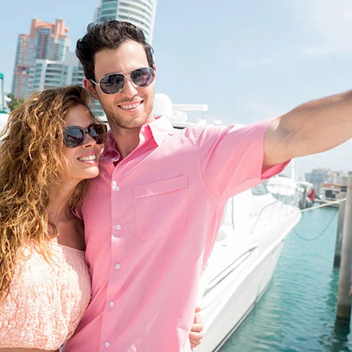 A couple is taking a selfie on a sunny day by a marina, with boats and city buildings in the background.