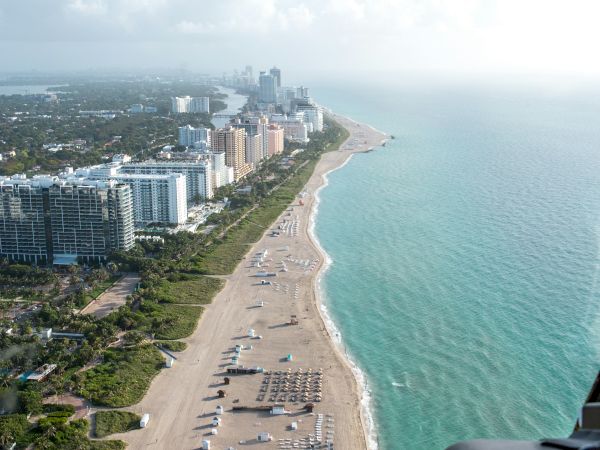 Aerial view of a beachfront cityscape with buildings, a sandy beach, and scattered beach umbrellas and loungers along the shoreline.