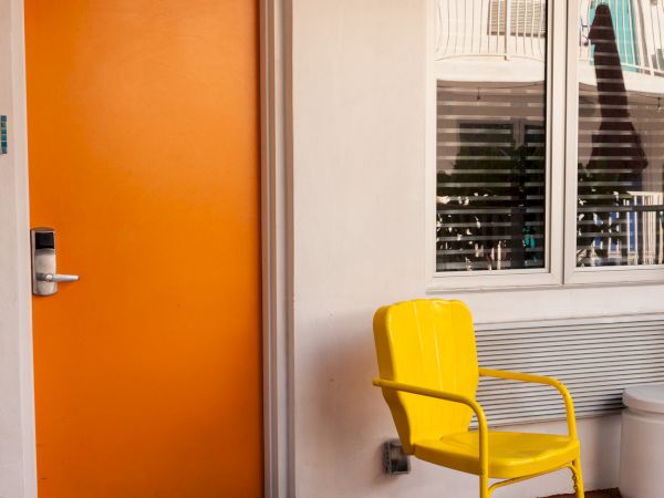 A bright orange door with a yellow chair nearby and a window reflecting a palm tree create a vibrant, modern outdoor space.