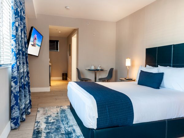 A neatly arranged hotel room with a bed, TV, blue decor, small dining area, and a window with patterned curtains on the side.