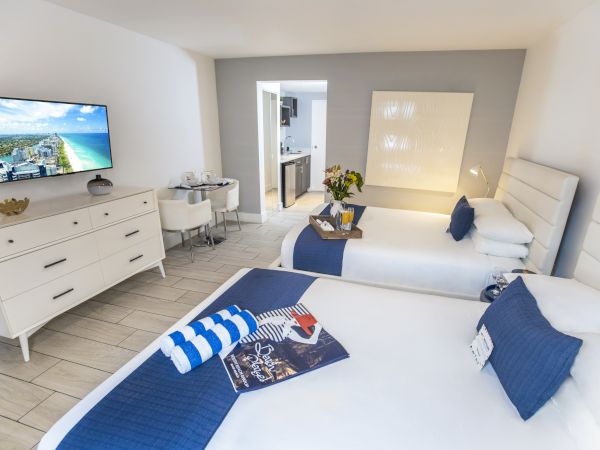 A modern hotel room with two beds, a TV, dresser, and small dining area; blue accents provide a nautical theme to the decor.
