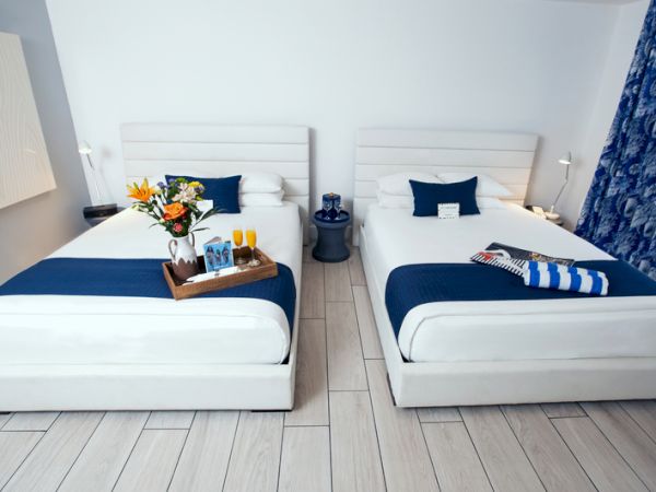 The image shows a room with two beds, blue accents, a tray with drinks and flowers, and a magazine placed on one of the beds.