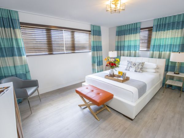 A modern bedroom with a bed, orange bench, side tables, lamps, and patterned curtains. A tray with flowers and items sits on the bed.