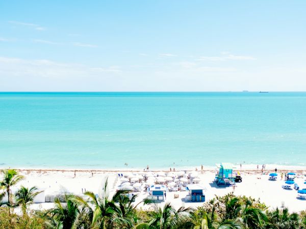 A pristine beach with turquoise waters, white sand, sun umbrellas, and palm trees under a clear blue sky, with people enjoying the scene.