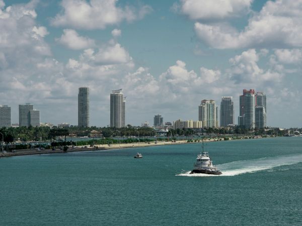 A small boat navigates a turquoise sea, heading towards a skyline of modern skyscrapers under a partly cloudy sky.