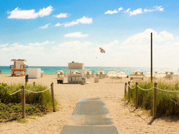 A picturesque beach scene with blue skies, parasailing, sun loungers, and a pathway leading to the water.