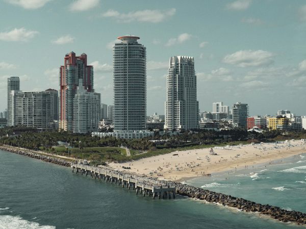 A beachfront cityscape with tall buildings, a pier extending into the water, and a sandy beach with people scattered across it.