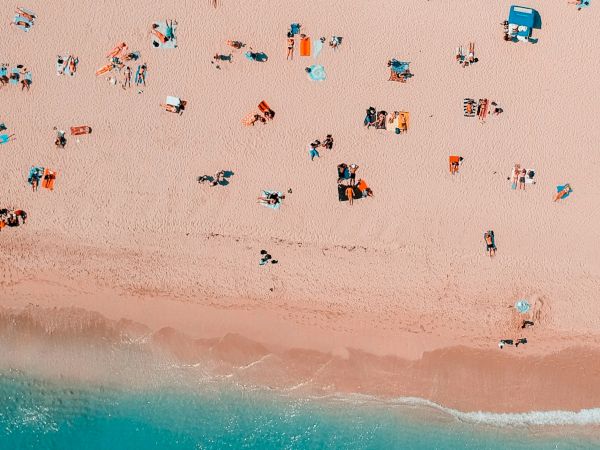 Aerial view of a beach features people, umbrellas, and towels on the sand with turquoise ocean waves.