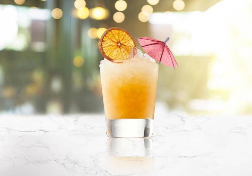 A refreshing cocktail with a gradient orange tint, garnished with a dried citrus wheel and a small pink umbrella on a marble surface.