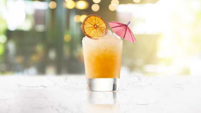 A refreshing cocktail with a gradient orange tint, garnished with a dried citrus wheel and a small pink umbrella on a marble surface.