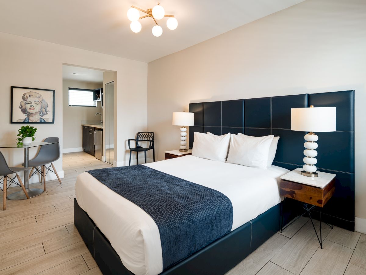 A modern hotel room with a large black upholstered bed, white linens and navy throw, nightstands with lamps, a small seating area, and neutral decor.