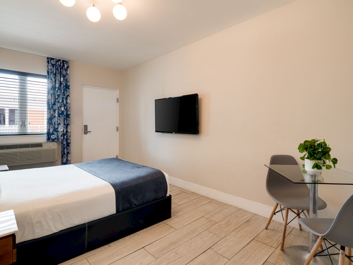 A clean hotel room with a double bed, wall-mounted TV, a small glass dining table and chairs, a window with blue patterned curtains, and a potted plant.