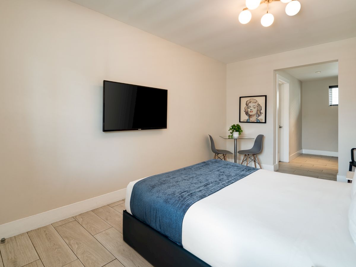 A modern hotel-style bedroom with a wall-mounted TV, a white bed with a blue throw, light wood floors, and a small desk area near a doorway.