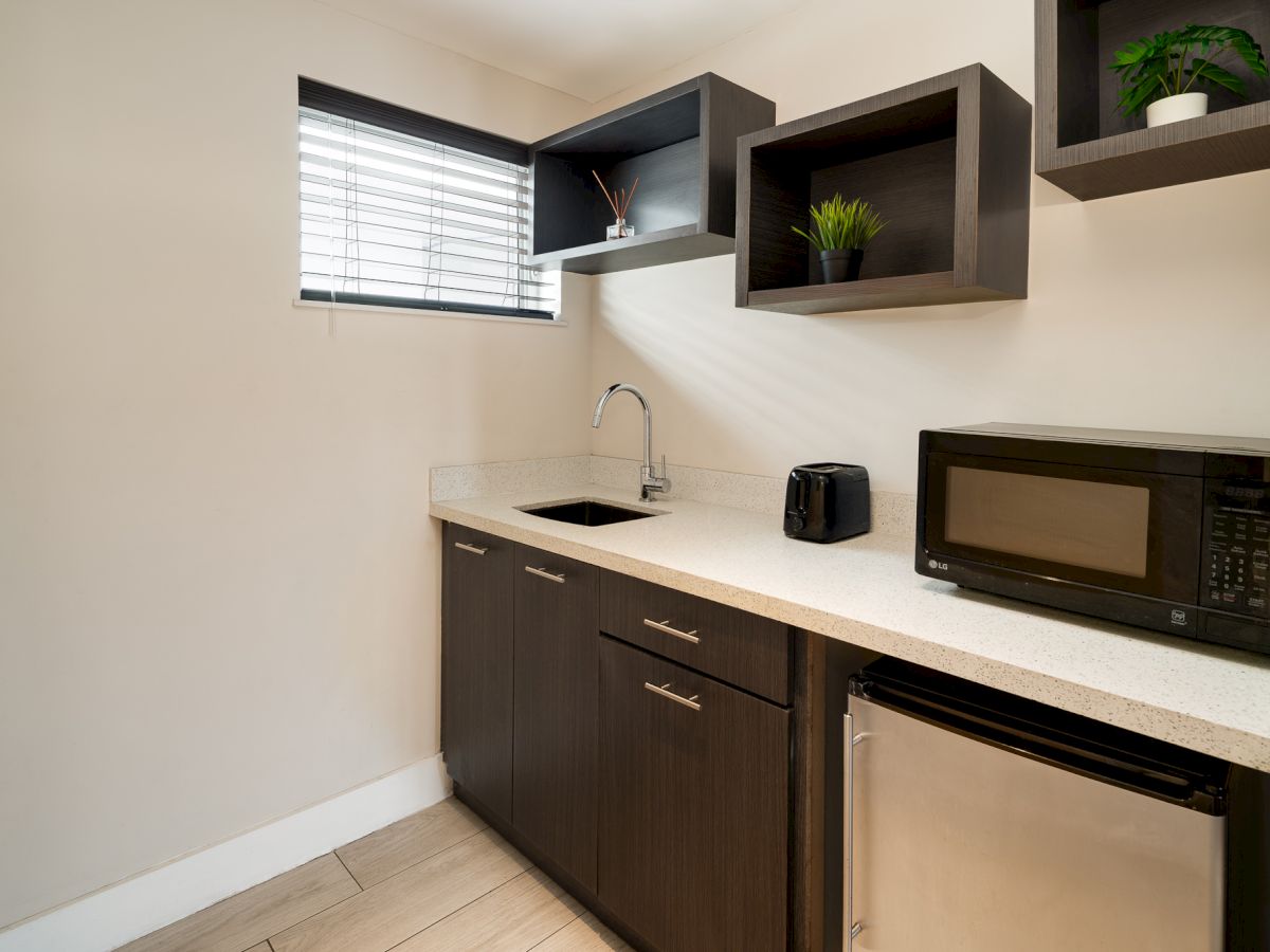 A compact kitchenette with dark cabinets, a white countertop, a small sink, a microwave, mini fridge, and floating shelves near a window.