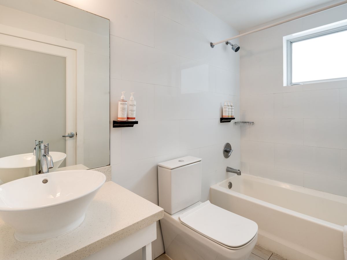 A modern bathroom with a white sink atop a vanity, a close-coupled toilet, a rectangular tub with a shower, glass shelves, and a small high window.