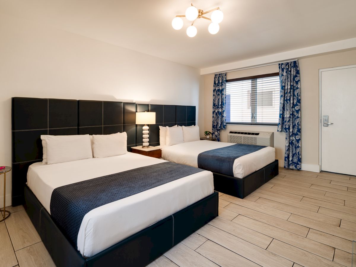 Two queen beds with dark headboards, nightstand lamp, window with blue curtains, and a clean, modern hotel room layout. ending sentence.