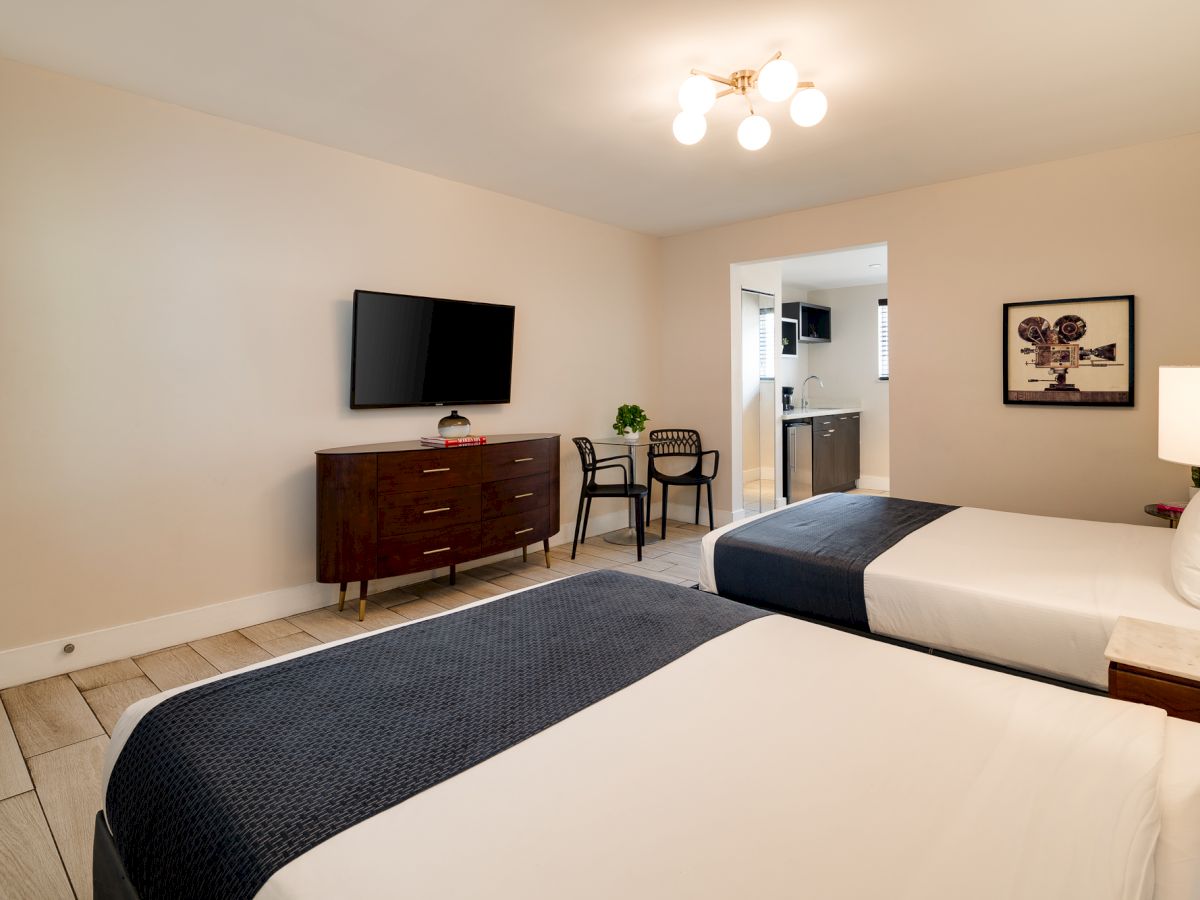 Two neatly made hotel beds with white linens and navy bed runners, a dresser with a wall TV, small table and chairs, and soft lighting.