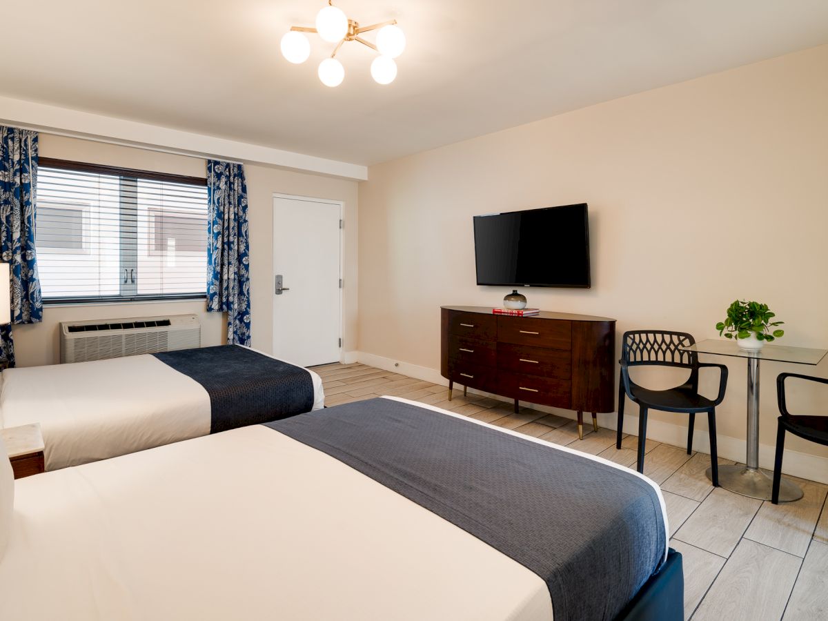 Two neatly made hotel beds with white linens and dark runners, a dresser with a wall-mounted TV, a small dining table with chairs, a window with blue curtains, and a bright ceiling light.