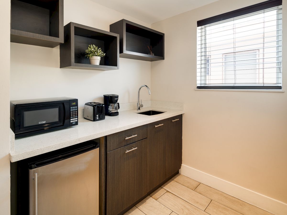 A compact kitchenette with dark wood cabinets, a white countertop, small fridge, microwave, toaster, coffee maker, sink, open shelves, and a window with blinds.