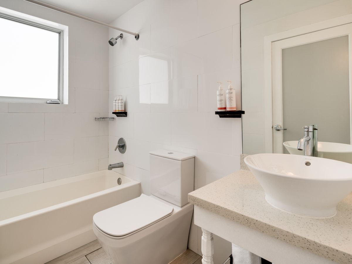 A clean bathroom with a tub, toilet, sink on a countertop, a mirror, and minimal decor with shelves and a window.
