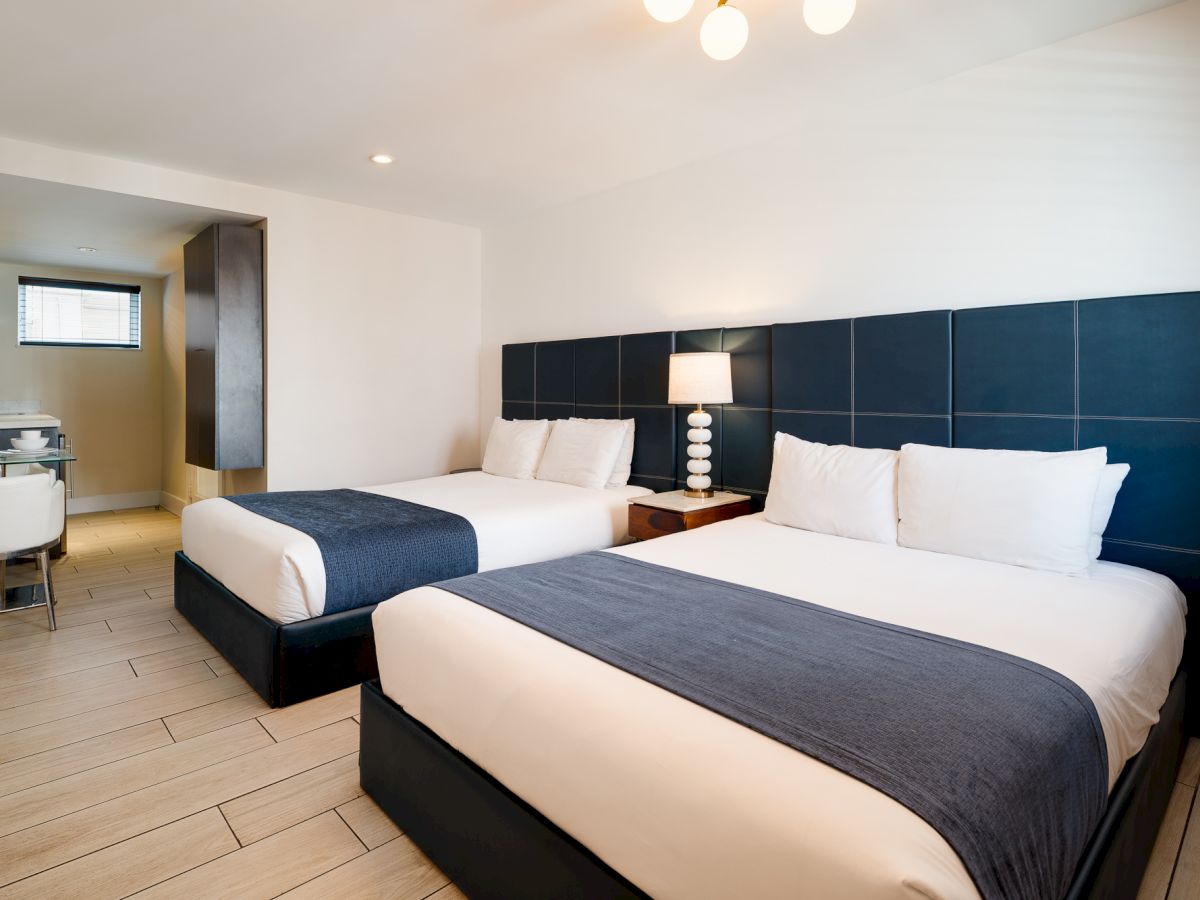 Two double beds with white linens and dark blue headboards, a bedside lamp, tiled floor, and a small kitchenette area in a modern hotel room.