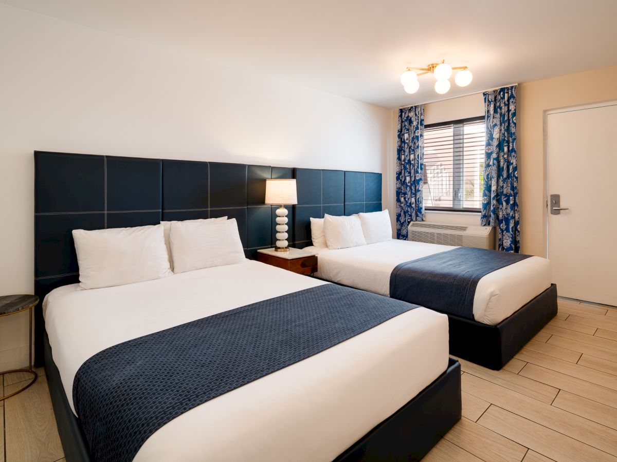 A clean hotel room with two single beds, dark blue headboards, white linens, navy throws, a small side table, a lamp, and a window with blue curtains.
