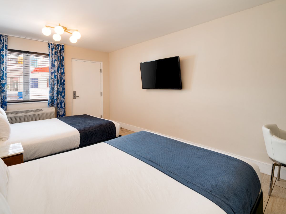 A bright hotel room with two single beds, blue throws, a wall-mounted TV, a small desk and chair, a window with blue curtains, and a door in the corner.