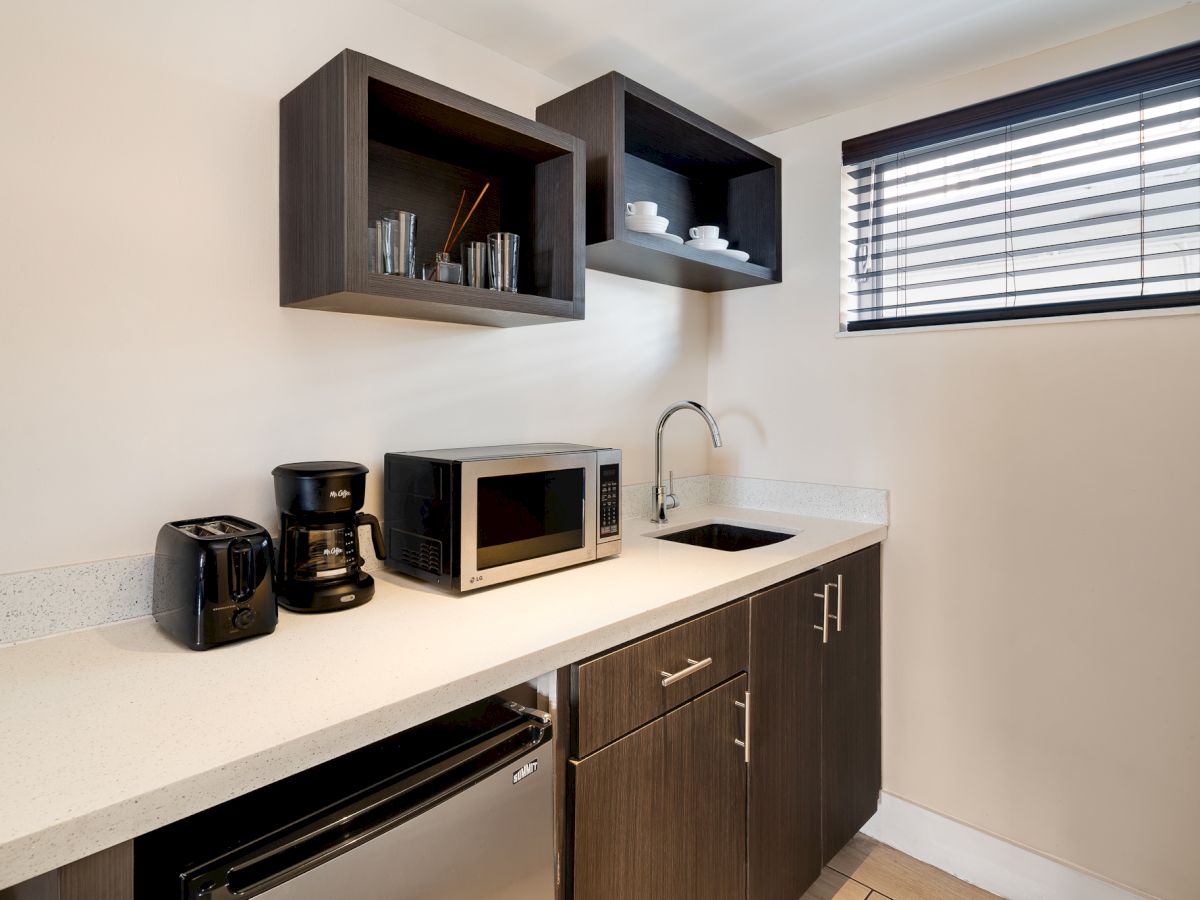 A compact kitchenette featuring dark wood cabinets, a white countertop, a microwave, coffee maker, toaster, sink, and a small window with blinds.