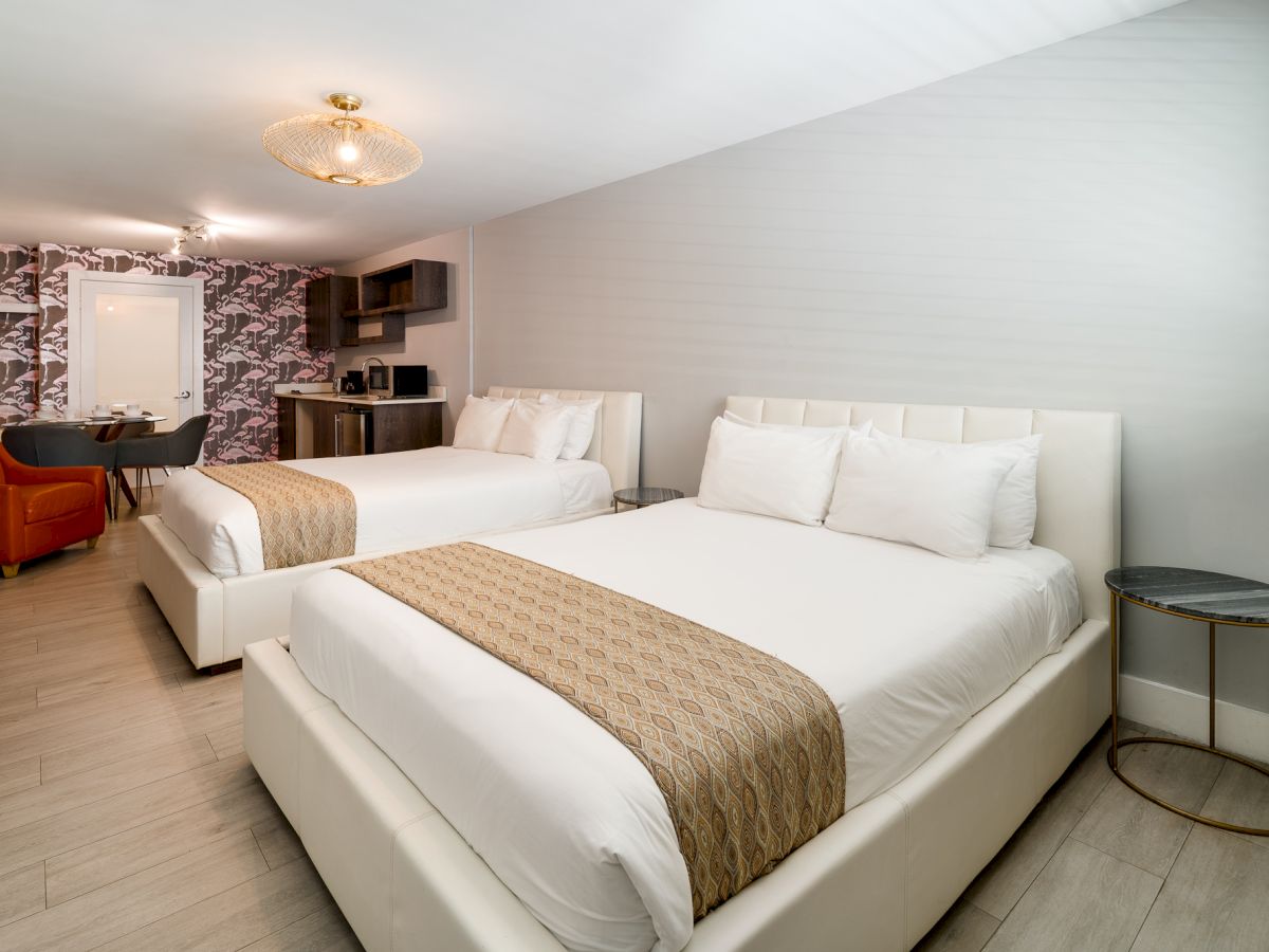 A bright hotel room with two white upholstered queen beds, tan runners, a small round side table, seating area, and a patterned accent wall in the background.