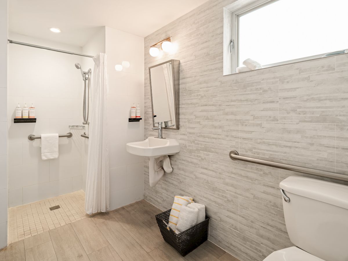 A modern bathroom with a walk-in shower, wall-mounted sink, mirror, window, toilet, towel bars, and a small bin of towels.