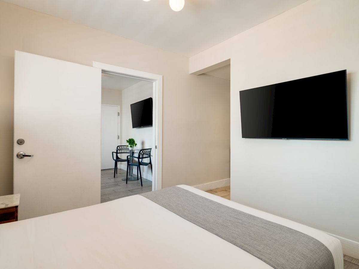A bright hotel room with a wall-mounted TV, white walls, a neatly made bed with a gray runner, and an open door revealing a small adjacent dining area.