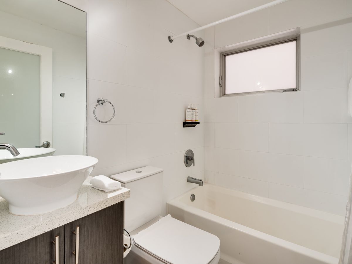 A modern bathroom with a white bathtub/shower, a pedestal sink on a dark vanity, a toilet, a small window, and simple fixtures.