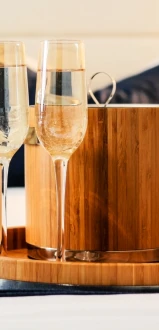 Two champagne flutes and an ice bucket with tongs sit on a round wooden tray, placed on a bed.