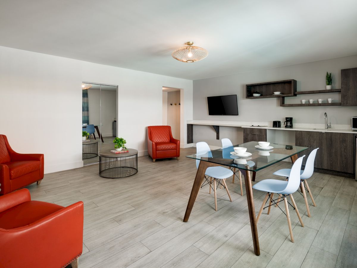 A modern, open-plan living and kitchen area with red armchairs, a dining table for six, wall-mounted TV, and light wood flooring.