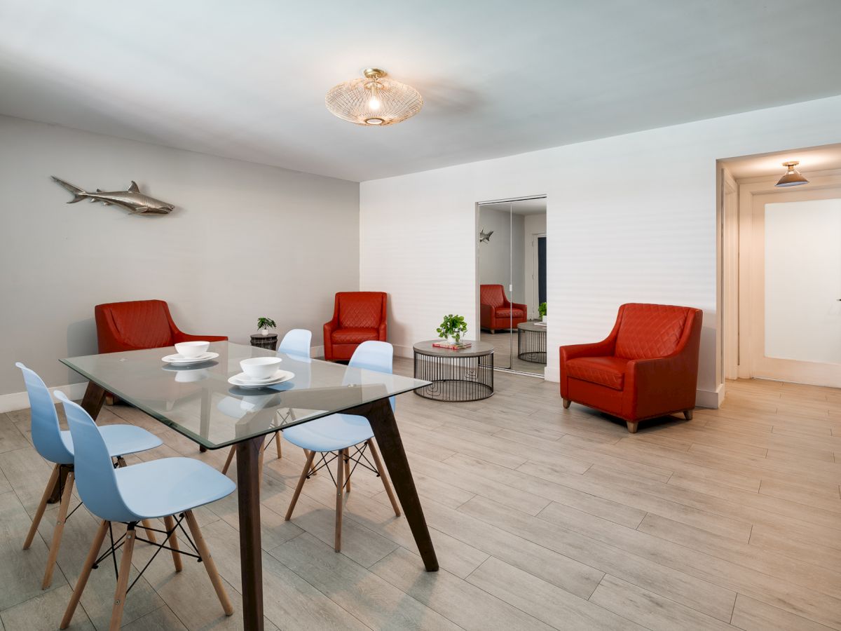 A bright, modern dining area with a glass table and pastel blue chairs, red armchairs, light wood floors, and a decor fish on the wall.