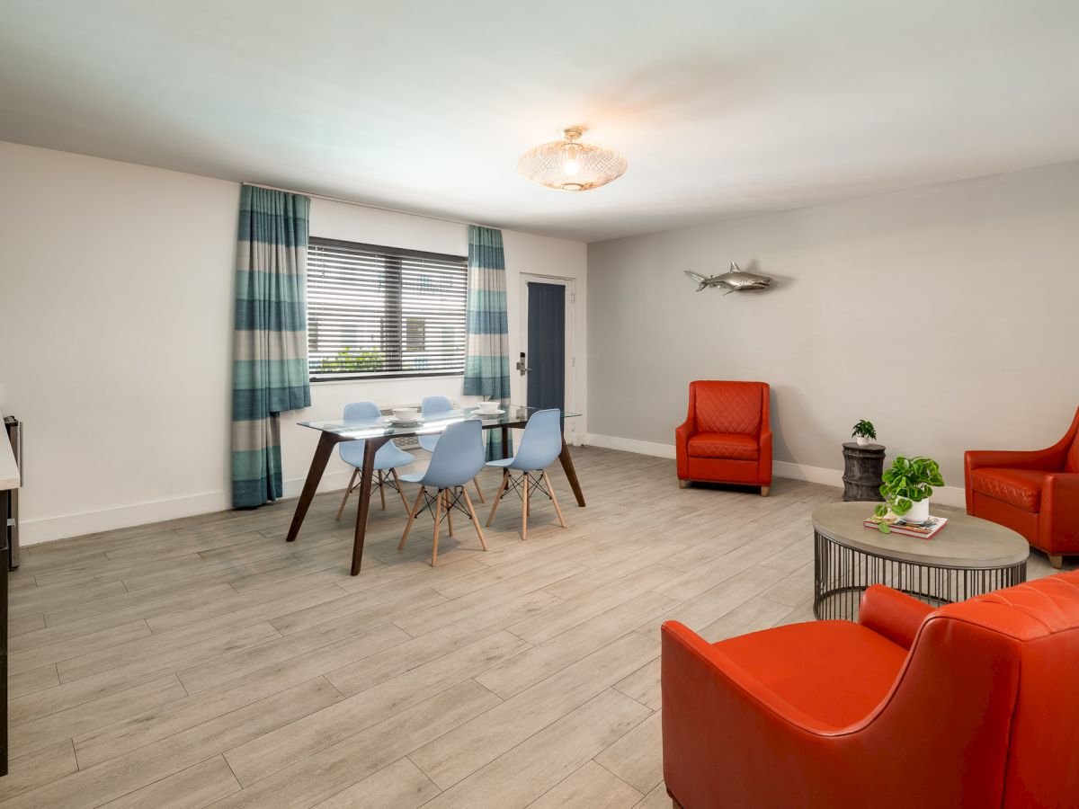 A bright, modern living room with light wood floors, a blue-and-white palette, a round dining table set, and bold orange chairs for contrast.