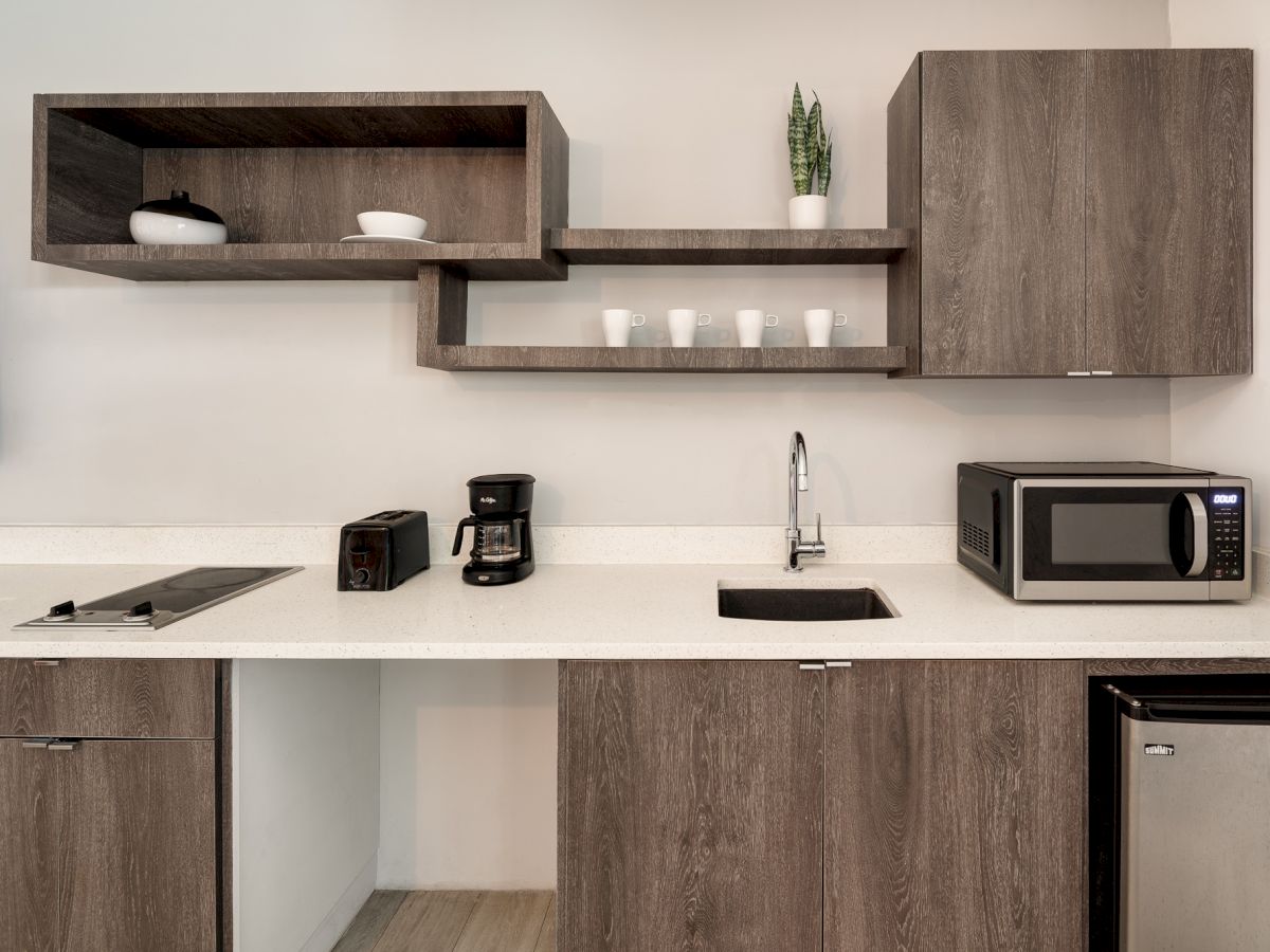 A modern kitchen with wooden cabinets, a white countertop, wall shelves, and small appliances like a toaster, coffee maker, microwave, and sink.