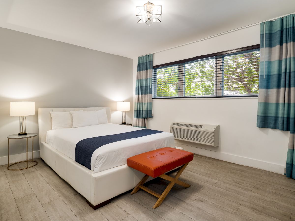 A clean, modern hotel room with a white bed, navy accents, two bedside lamps, a red ottoman at the foot, striped blue curtains, and a window with an air conditioner.