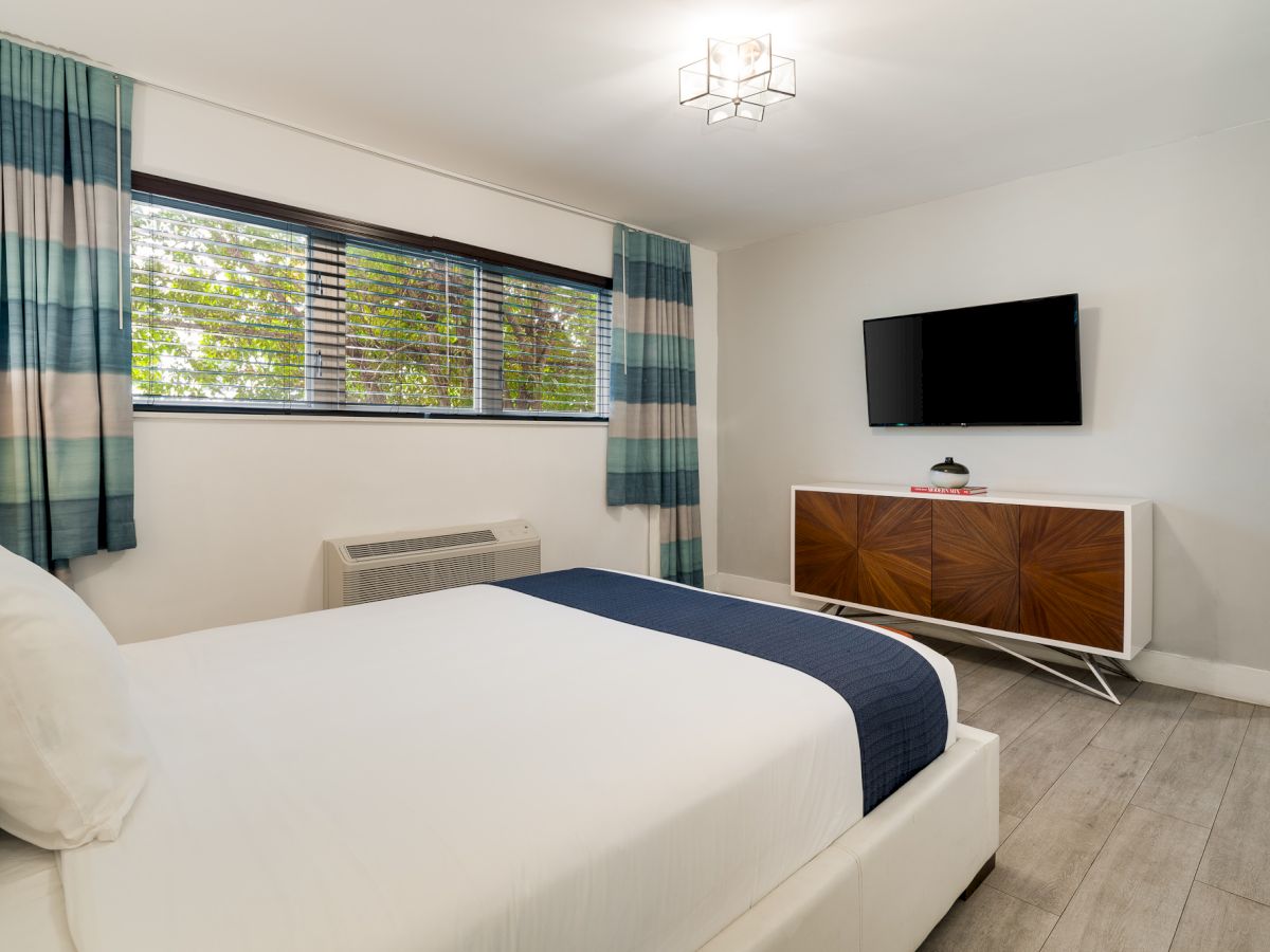 A modern hotel bedroom with a white bed, blue-gray curtains, a wall-mounted TV, and a wooden dresser, bright and airy.