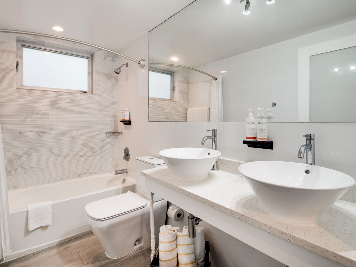 A modern bathroom with a marble-tiled tub area, a glass shower, dual vessel sinks on a light countertop, a toilet, large mirror, and neatly stacked towels.