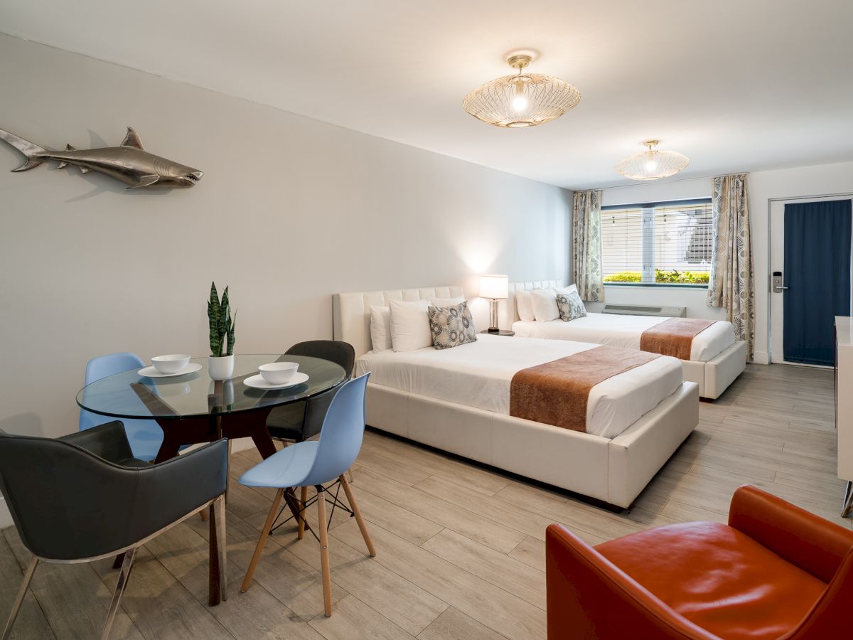 A bright, modern hotel suite with two white beds, a small dining table for four, a red chair, a blue door, and a decorative shark on the wall.