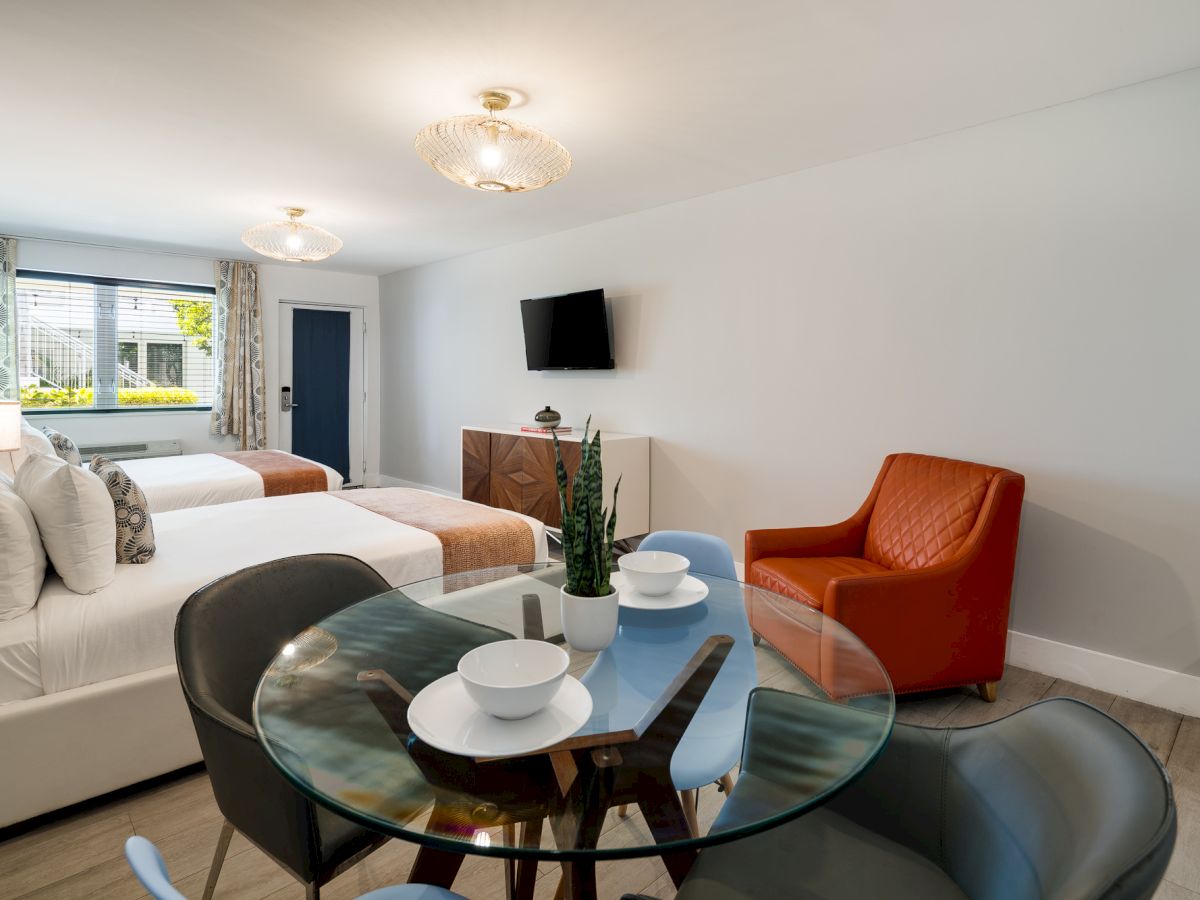 A bright hotel suite with a white sofa, glass dining table, two chairs, an orange armchair, flat-screen TV, and a view of the window; modern decor.